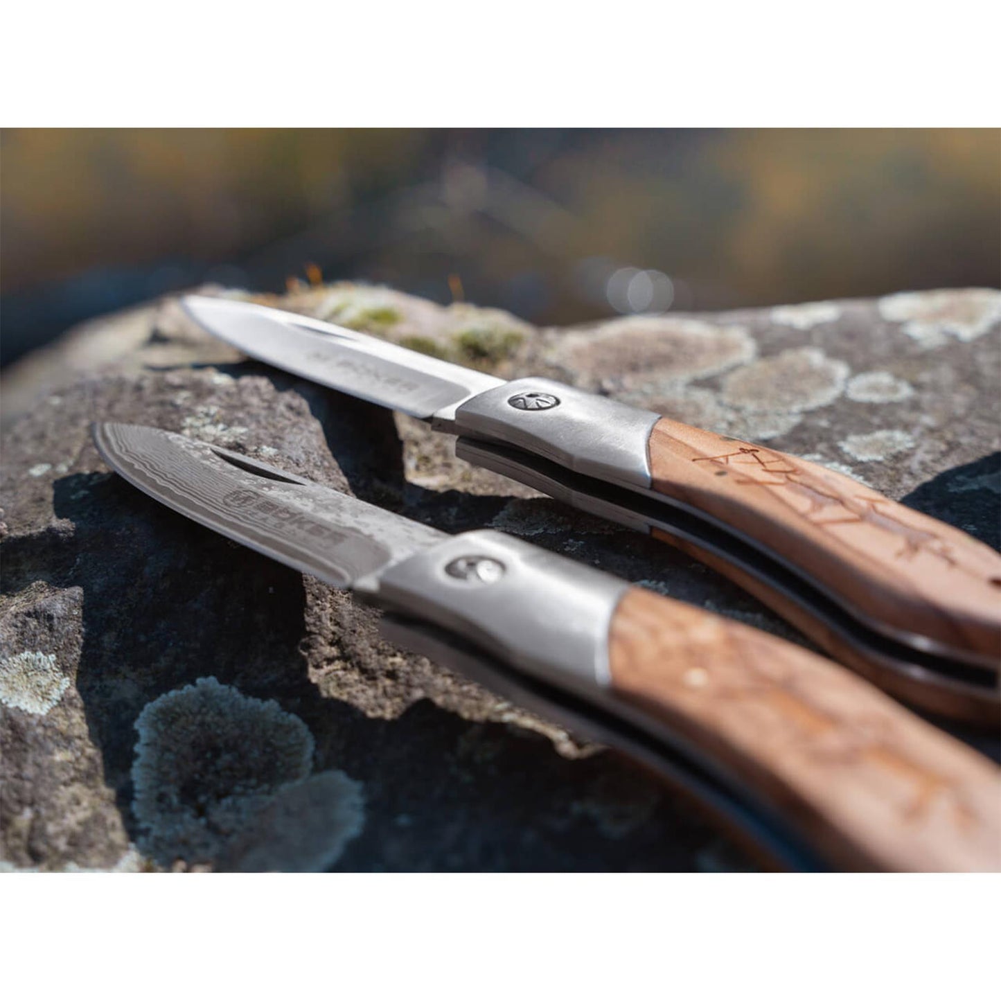 Boker Magnum Caveman sulankstomas peilis su medine rankena ir dekoruota geležte, padetas ant akmens pavirsiaus
