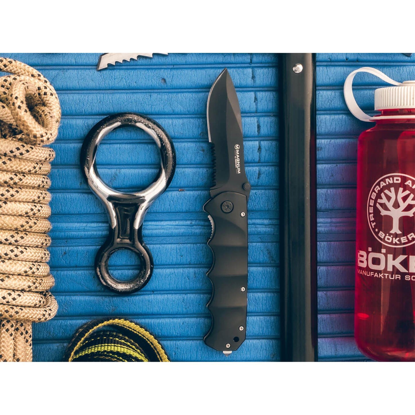 Boker Black Spear sulenkiamas peilis su juoda geležte ir aliuminio rankena, rodomas ant melynos teksturuotos atramos